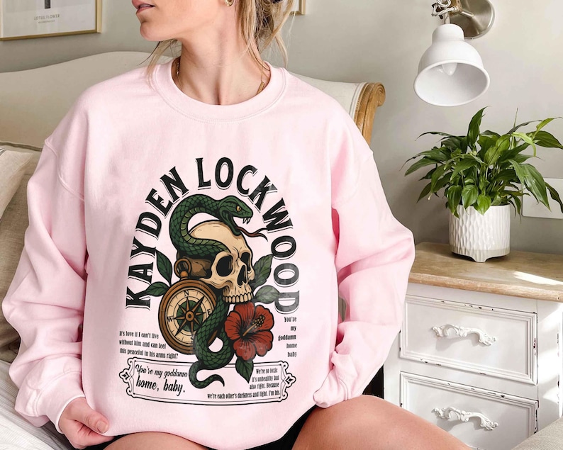 May include: A light pink sweatshirt with the text "KAYDEN LOCKWOOD" in a curved arch. The graphic includes a skull, a green snake, a compass, and a red flower. Additional text is below the graphic.