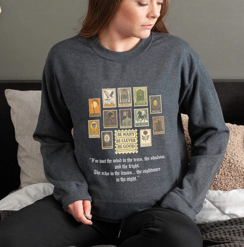 May include: Dark gray sweatshirt featuring a collection of vintage-style postage stamps. The stamps are arranged in a square and include various illustrations. Below the stamps is the text "BE WARY BE CLEVER BE GOOD" and a quote about the wind and the night.