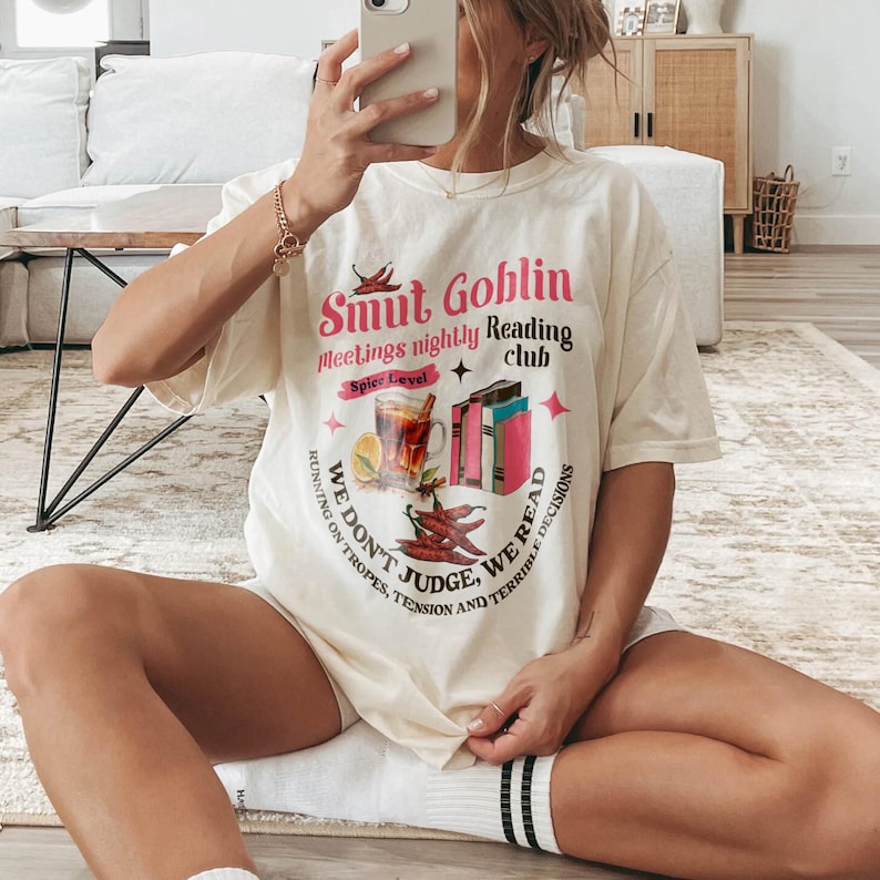 May include: Cream-colored graphic tee with the text "Smut Goblin Meetings nightly Reading club". The design includes illustrations of books, chili peppers, and a drink. The shirt has a relaxed fit.