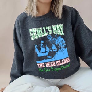 May include: Dark gray sweatshirt with a graphic featuring the text "SKULL'S BAY" above a blue illustration of a ship, island, and palm trees. Below the image is the text "THE DEAD ISLANDS The Sea Dragon (Tavern)".