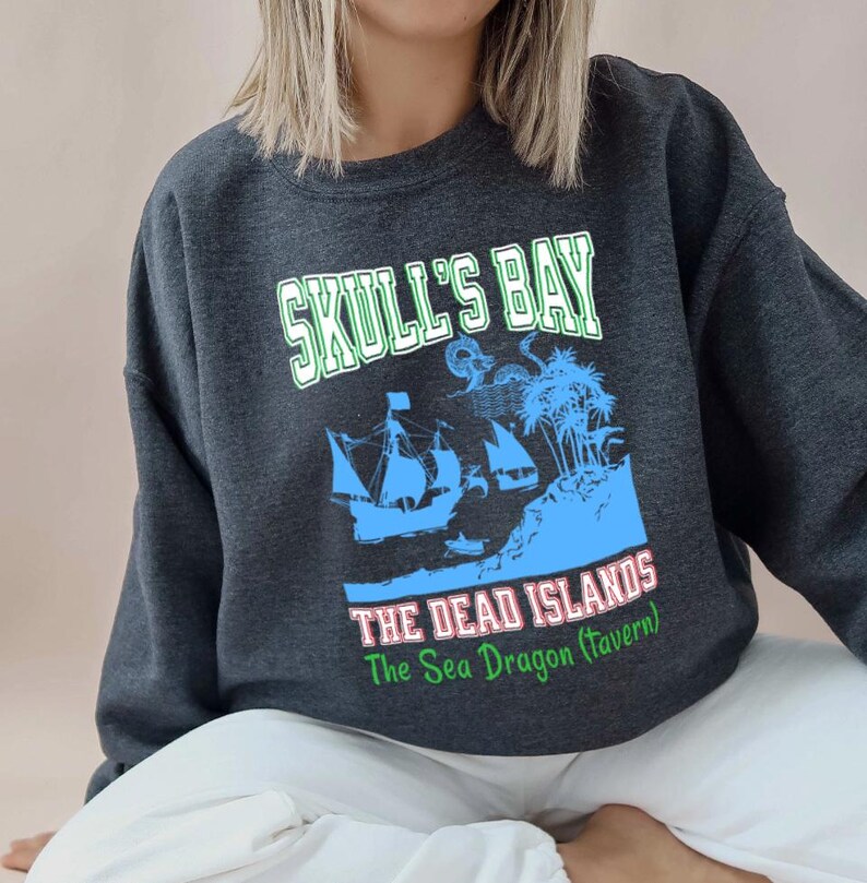 May include: Dark gray sweatshirt with a graphic featuring the text "SKULL'S BAY" above a blue illustration of a ship, island, and palm trees. Below the image is the text "THE DEAD ISLANDS The Sea Dragon (Tavern)".