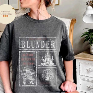 Op de afbeelding: Een donkergrijs T-shirt met een witte grafische print die "Welkom bij de Blunder" zegt met drie kleinere vakjes eronder die zeggen "Wees voorzichtig, Wees slim, Wees goed". De grafiek bevat ook een kasteel, een boom en een stapel boeken.