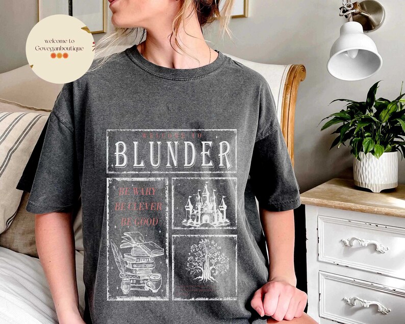 Op de afbeelding: Een donkergrijs T-shirt met een witte grafische print die "Welkom bij de Blunder" zegt met drie kleinere vakjes eronder die zeggen "Wees voorzichtig, Wees slim, Wees goed". De grafiek bevat ook een kasteel, een boom en een stapel boeken.