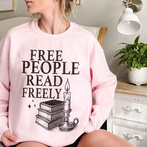 Op de afbeelding: Een lichtroze sweatshirt met de zwarte tekst "FREE PEOPLE READ FREELY". Daaronder een afbeelding van een stapel boeken en een brandende kaars. De sweatshirt heeft een ronde hals.