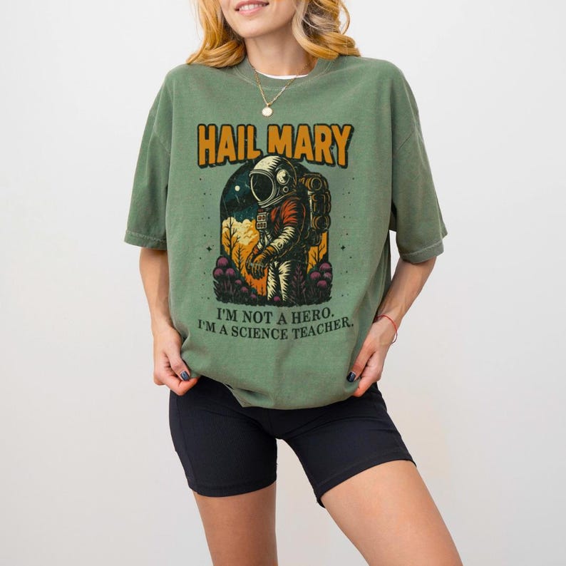 May include: Olive green t-shirt with the text "HAIL MARY" above an astronaut graphic. Below the graphic, the text reads "I'M NOT A HERO. I'M A SCIENCE TEACHER."