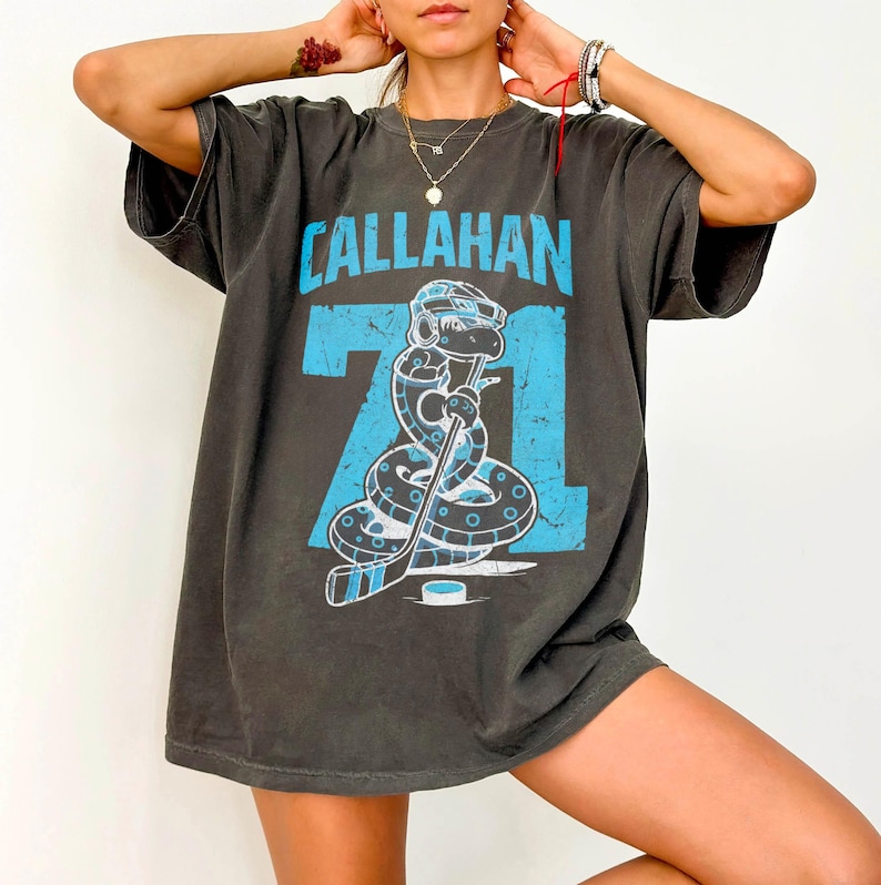 May include: Dark gray t-shirt with the name "CALLAHAN" in light blue block letters above the number "71". The graphic features a snake wearing a hockey helmet and holding a hockey stick. The shirt is oversized.