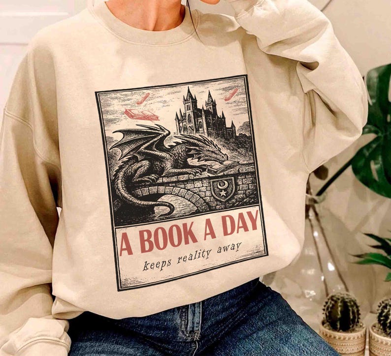 May include: Beige sweatshirt featuring a black and white illustration of a dragon, castle, and the text "A BOOK A DAY keeps reality away." The graphic design is in a vintage style. The sweatshirt is a casual, comfortable garment.