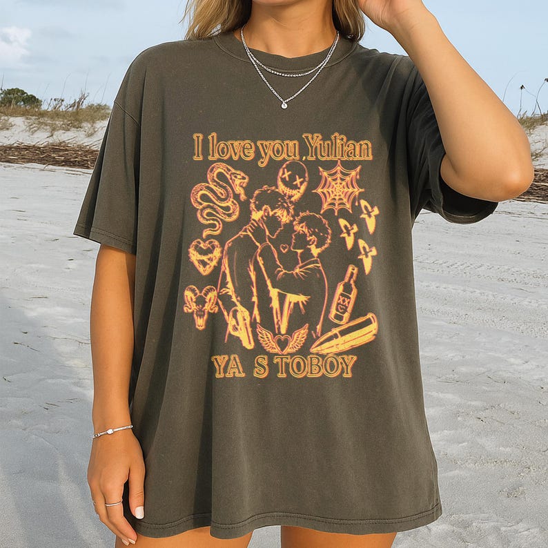 May include: Dark olive green t-shirt with a graphic design in shades of orange and yellow. The design includes the text "I love you Yulian" and "YA S TOBOY", along with illustrations of a snake, a spiderweb, and two figures kissing.