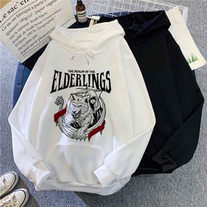 May include: White hoodie with a front pocket and drawstring. The graphic on the front features a wolf, the text "THE REALM OF THE ELDERLINGS", and other design elements. A black hoodie is next to it.