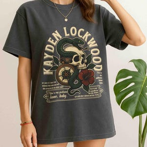 May include: Dark gray t-shirt featuring a graphic design with the text "KAYDEN LOCKWOOD". The design includes a skull, snake, compass, and a red hibiscus flower. The shirt has short sleeves and a crew neck.