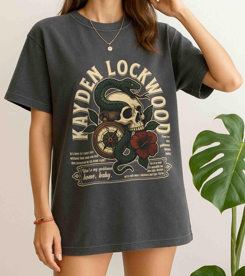 May include: Dark gray t-shirt featuring a graphic design with the text "KAYDEN LOCKWOOD". The design includes a skull, snake, compass, and a red hibiscus flower. The shirt has short sleeves and a crew neck.