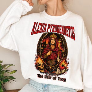 Op de afbeelding: Witte sweatshirt met een afbeelding van een vrouw in een rode jurk en kroon, met een kruis. De tekst "ALEXIA PYROGENNETOS" staat in het rood boven de afbeelding en "The Heir of Troy" eronder. Een schedel en een vlammend hart zijn ook aanwezig.