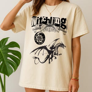 May include: Cream-coloured t-shirt with the word "Witchling" in black above a graphic of roses, swords, a rose, a crescent moon, and a dragon. The dragon is black with outstretched wings.