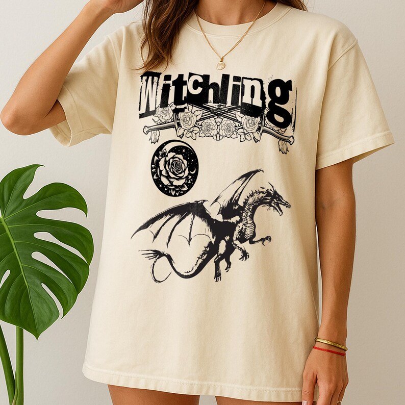 May include: Cream-coloured t-shirt with the word "Witchling" in black above a graphic of roses, swords, a rose, a crescent moon, and a dragon. The dragon is black with outstretched wings.