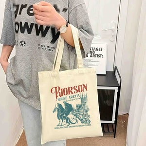 May include: A beige tote bag with a graphic design featuring a blue dragon and a castle. The text on the bag reads "Riorson House Aretia Rebellion & Regia Welcome Rebel Faction Basgiath War College".