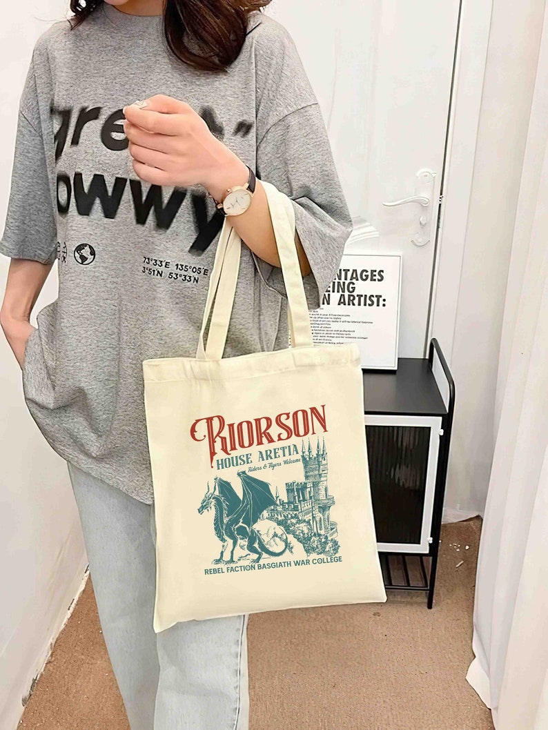 May include: A beige tote bag with a graphic design featuring a blue dragon and a castle. The text on the bag reads "Riorson House Aretia Rebellion & Regia Welcome Rebel Faction Basgiath War College".
