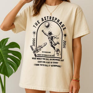 May include: Cream-colored t-shirt featuring a black graphic design. The design includes an astronaut, a sun, a spaceship, and the text "THE ASTROPHAGE." Additional text reads "THE SILVER DUST, ENERGY DEVOURER OF THE STARS." The shirt also has the text "WHAT WOULD YOU CALL AN ORGANISM THAT EXISTS ON A DIET OF STARS? I THINK YOU'D CALL IT 'ASTROPHAGE.'"