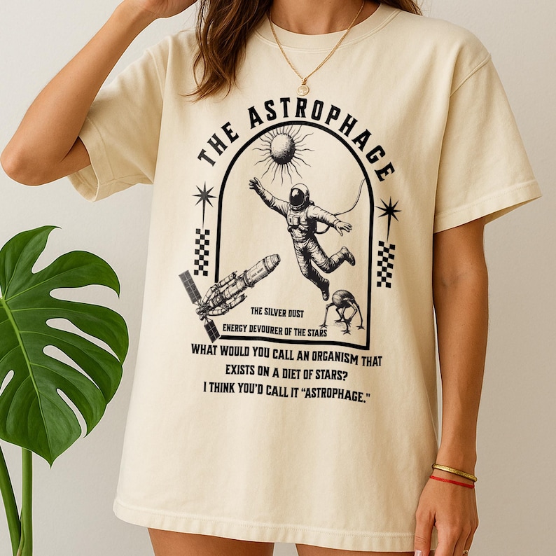 May include: Cream-colored t-shirt featuring a black graphic design. The design includes an astronaut, a sun, a spaceship, and the text "THE ASTROPHAGE." Additional text reads "THE SILVER DUST, ENERGY DEVOURER OF THE STARS." The shirt also has the text "WHAT WOULD YOU CALL AN ORGANISM THAT EXISTS ON A DIET OF STARS? I THINK YOU'D CALL IT 'ASTROPHAGE.'"