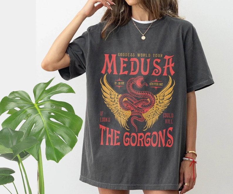 K&ouml;nnte beinhalten: Ein dunkelgraues T-Shirt mit einem grafischen Druck von Medusa, einer mythischen griechischen Figur mit Schlangenhaaren. Der Text "Goddess World Tour" und "The Gorgons" ist auf dem Shirt gedruckt.