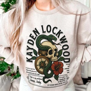 May include: Beige sweatshirt with a graphic design featuring a skull, snake, compass, and red hibiscus flower. The text reads "KAYDEN LOCKWOOD" and includes phrases like "You're my goddamn home, baby."