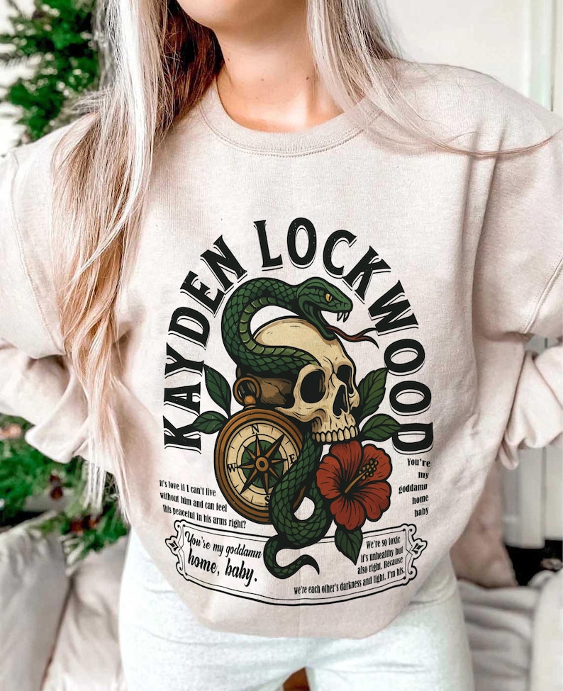 May include: Beige sweatshirt with a graphic design featuring a skull, snake, compass, and red hibiscus flower. The text reads "KAYDEN LOCKWOOD" and includes phrases like "You're my goddamn home, baby."