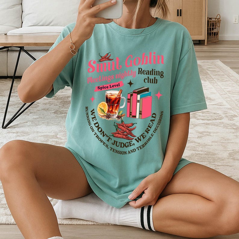 May include: A teal-colored t-shirt with the text "Smut Goblin Reading Club" and illustrations of books, chili peppers, and a drink. The shirt also includes the phrases "We don't judge, we read" and "Running on tropes, tension and terrible decisions."