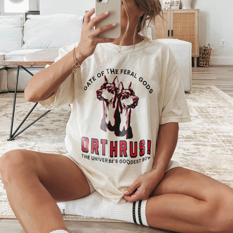 Puede incluir: Camiseta color crema con un gr&aacute;fico de perros de dos cabezas y el texto "GATE OF THE FERAL GODS" encima de la imagen. Debajo de la imagen, el texto dice "ORTHRUS! THE UNIVERSE'S GOODEST BOY". La camiseta se usa de forma informal.