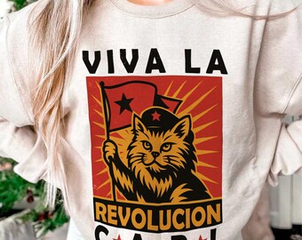 May include: Beige sweatshirt featuring a graphic of a cat holding a flag with a star, set against a red and yellow background. The text "VIVA LA REVOLUCION C*A*R*L" is printed above and below the graphic in black.