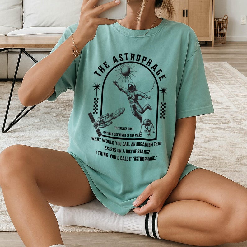 May include: A teal-colored t-shirt with the text "THE ASTROPHAGE" and an illustration of an astronaut, a rocket, and a creature. The text below reads "THE SILVER DUST ENERGY DEVOURER OF THE STARS".