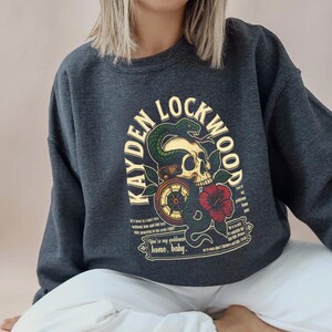 May include: Dark gray sweatshirt featuring a skull with a snake, compass, and red flower graphic. The text "KAYDEN LOCKWOOD" arches above the skull. Additional text includes phrases like "home, baby" and other quotes.