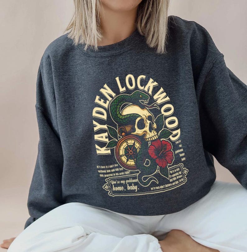 May include: Dark gray sweatshirt featuring a skull with a snake, compass, and red flower graphic. The text "KAYDEN LOCKWOOD" arches above the skull. Additional text includes phrases like "home, baby" and other quotes.