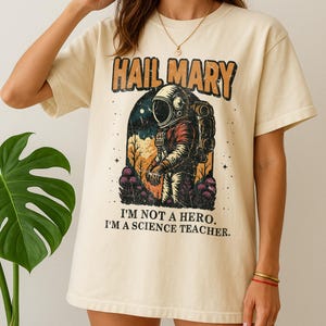 May include: Cream-colored t-shirt featuring a retro graphic of an astronaut within an archway, with the text "HAIL MARY" above and "I'M NOT A HERO. I'M A SCIENCE TEACHER." below. The astronaut is wearing a red and orange spacesuit.