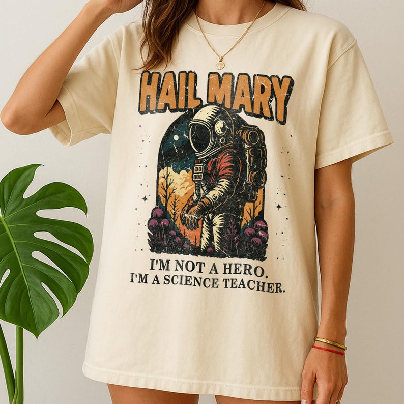 May include: Cream-colored t-shirt featuring a retro graphic of an astronaut within an archway, with the text "HAIL MARY" above and "I'M NOT A HERO. I'M A SCIENCE TEACHER." below. The astronaut is wearing a red and orange spacesuit.