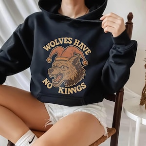 May include: Black hooded sweatshirt with the text "WOLVES HAVE NO KINGS" and a graphic of a wolf wearing a jester's hat. The design is in shades of brown and tan.