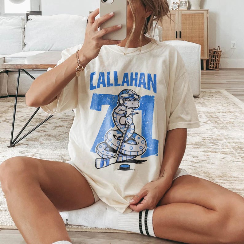 May include: Cream-colored t-shirt with the word "CALLAHAN" in blue block letters above a blue number "71" and a cartoon snake wearing a hockey helmet and holding a hockey stick. The snake is on a hockey puck.