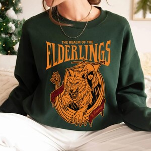 May include: Dark green sweatshirt with an orange graphic. The graphic features a wolf, a hooded figure, and the text "THE REALM OF THE ELDERLINGS". A banner with text is below the wolf. The sweatshirt is a crew neck style.
