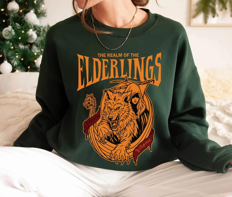May include: Dark green sweatshirt with an orange graphic. The graphic features a wolf, a hooded figure, and the text "THE REALM OF THE ELDERLINGS". A banner with text is below the wolf. The sweatshirt is a crew neck style.