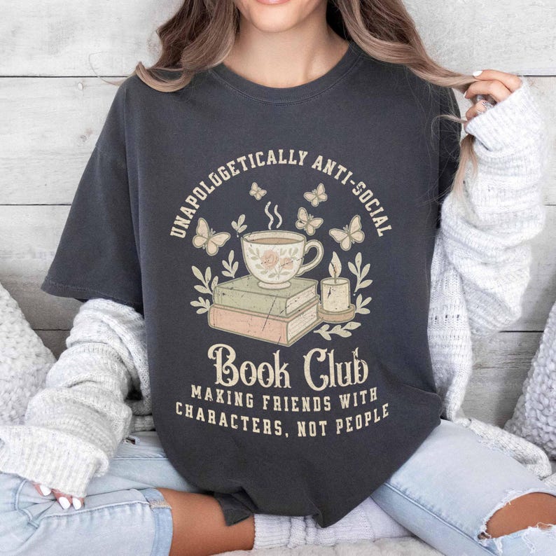 May include: Dark gray t-shirt with a graphic design featuring a teacup, books, butterflies, and a candle. The text reads "UNAPOLOGETICALLY ANTI-SOCIAL" and "Book Club MAKING FRIENDS WITH CHARACTERS, NOT PEOPLE."