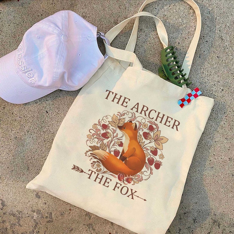 May include: A beige canvas tote bag with a fox illustration and the text "The Archer The Fox".