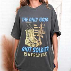 May include: Dark gray t-shirt with a graphic design. The design features a combat boot, a rifle, and an American flag, with the text "THE ONLY GOOD RIOT SOLDIER IS A DEAD ONE" in blue and yellow.