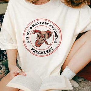 May include: Cream-colored t-shirt featuring a circular graphic with a skull and a red snake. The text around the graphic reads "ARE YOU GOING TO BE MY FOREVER FRECKLES?" The shirt is a casual style.