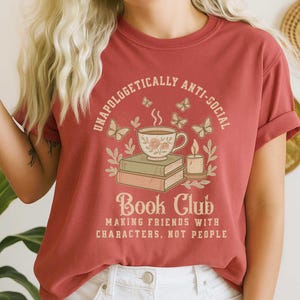 May include: A rust-colored t-shirt with a graphic design featuring a teacup, books, butterflies, and a candle. The text reads "Unapologetically Anti-Social" and "Book Club: Making Friends with Characters, Not People."