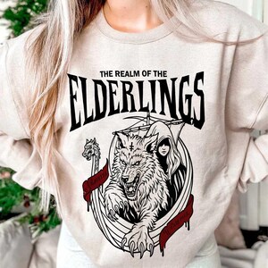 May include: Beige sweatshirt with a black and red graphic. The graphic features a wolf, a woman, and the text "THE REALM OF THE ELDERLINGS". A banner reads "WOLF PACK".