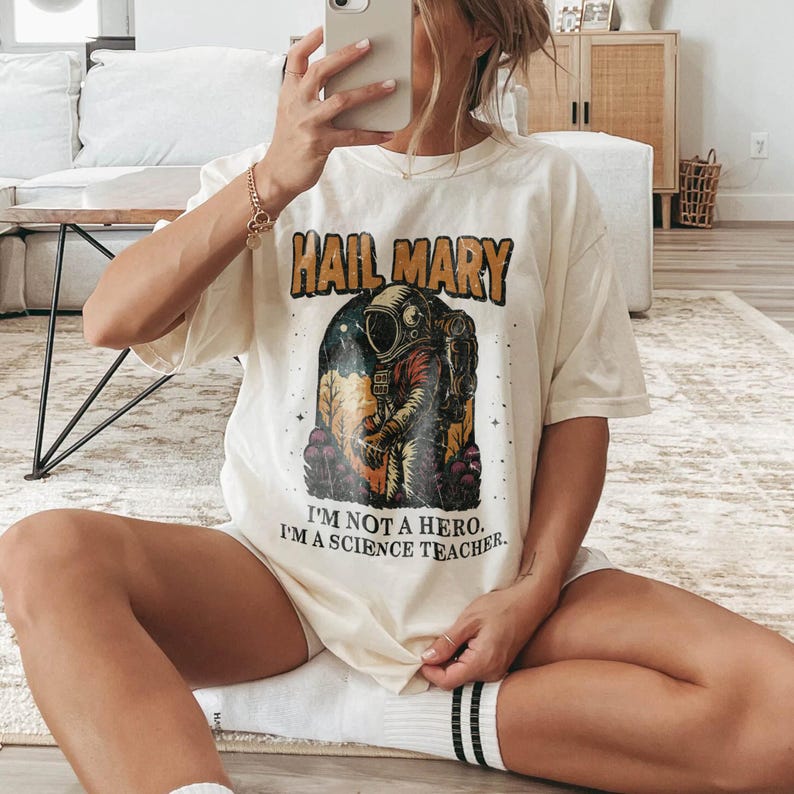 May include: Cream-colored t-shirt with the text "HAIL MARY" above an astronaut graphic. Below the graphic, the text reads "I'M NOT A HERO. I'M A SCIENCE TEACHER." The shirt has short sleeves and a relaxed fit.