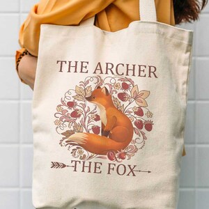 May include: A beige canvas tote bag with a fox illustration and the text "The Archer The Fox".