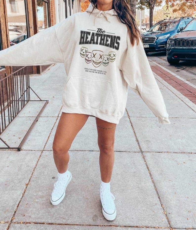 May include: Cream-coloured hoodie featuring the text "The Heathens" in black above skull faces. The hoodie has a front pocket and a drawstring hood. The person is wearing white trainers.