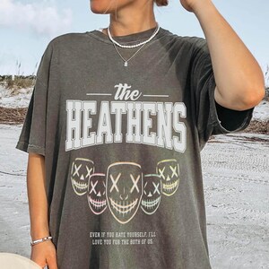 May include: Dark gray t-shirt with the text "The Heathens" in white block letters. Below the text are five stylized mask graphics with the text "XX" and a smiling mouth. The bottom of the shirt reads "EVEN IF YOU HATE YOURSELF, I'LL LOVE YOU FOR THE BOTH OF US."