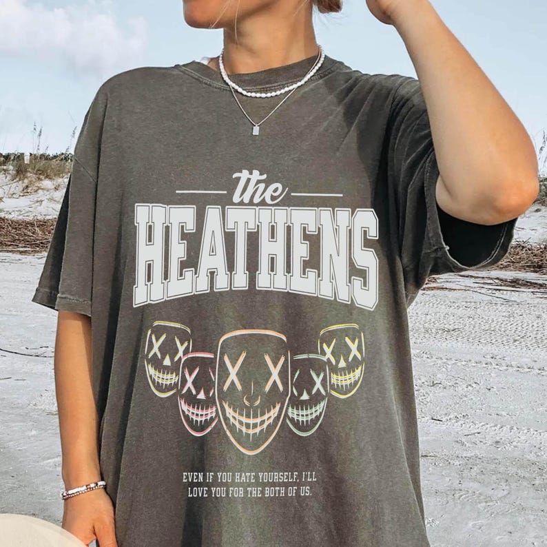 May include: Dark gray t-shirt with the text "The Heathens" in white block letters. Below the text are five stylized mask graphics with the text "XX" and a smiling mouth. The bottom of the shirt reads "EVEN IF YOU HATE YOURSELF, I'LL LOVE YOU FOR THE BOTH OF US."