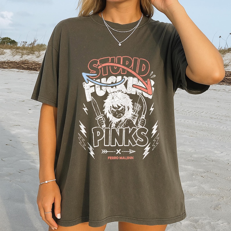 May include: Dark olive green t-shirt with a graphic design. The design features the words "STUPID FUCKING PINKS" in a stylized font, along with an illustration of a character holding knives and arrows. The text "FERRO MALJNN" is below the graphic.