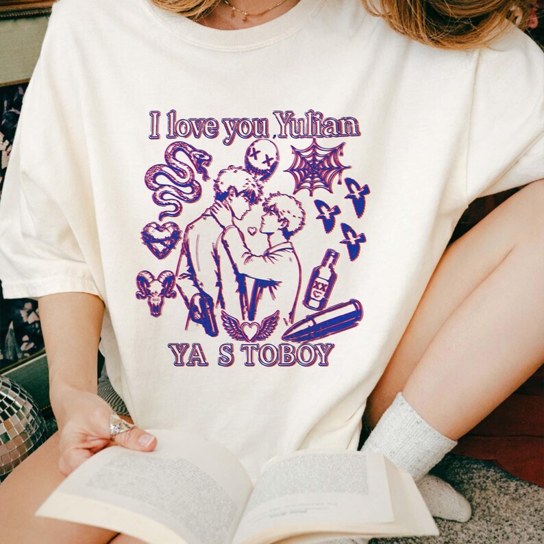 May include: Cream-colored t-shirt with a purple graphic design. The design includes the text "I love you Yulian" and "YA S TOBOY", along with illustrations of two men, a snake, a spiderweb, and other symbols.